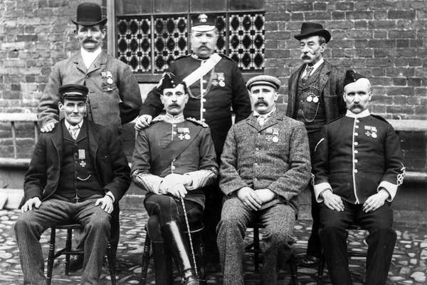 Posed black and white photo of seven men, four on chairs and three standing behind, wearing medals.