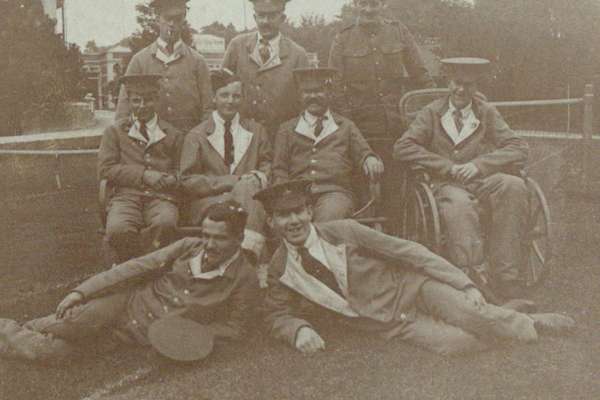 A group of First World War soldiers posing for a photo on a tennis court. Three sit on a bench, three stand behind, two lie on the floor and one is using a wheelchair.