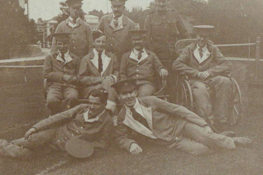 A group of First World War soldiers posing for a photo on a tennis court. Three sit on a bench, three stand behind, two lie on the floor and one is using a wheelchair.
