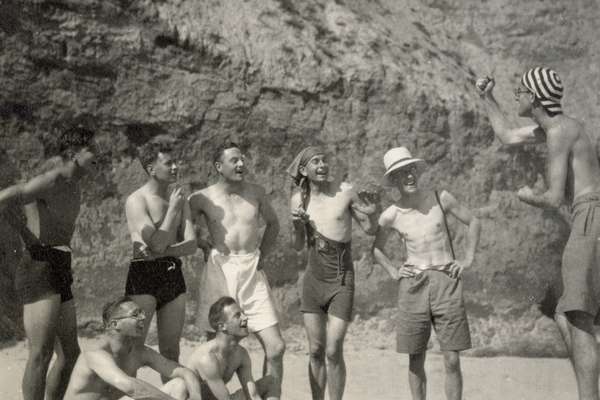 Monochrome photograph of a group of men in swimsuits on a beach. One of them is speaking jokingly from a box with a raised fist and the others look and him while laughing.