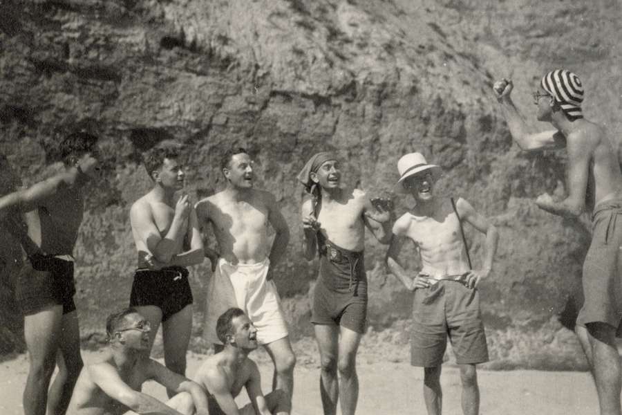 Monochrome photograph of a group of men in swimsuits on a beach. One of them is speaking jokingly from a box with a raised fist and the others look and him while laughing.