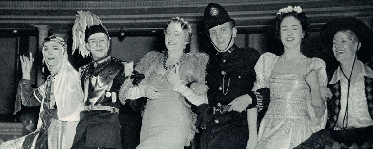 Six partygoers dressed elaborately lock arms and smile as they kick a leg up in unison.