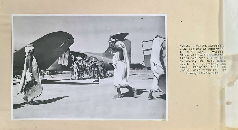 A typewritten caption beside a black and white photograph of an airfield scene. About ten men in uniform are loading a vehicle into a military plane. Manipuri women with bare feet and loose dress are crossing the airfield carrying baskets on their heads.
