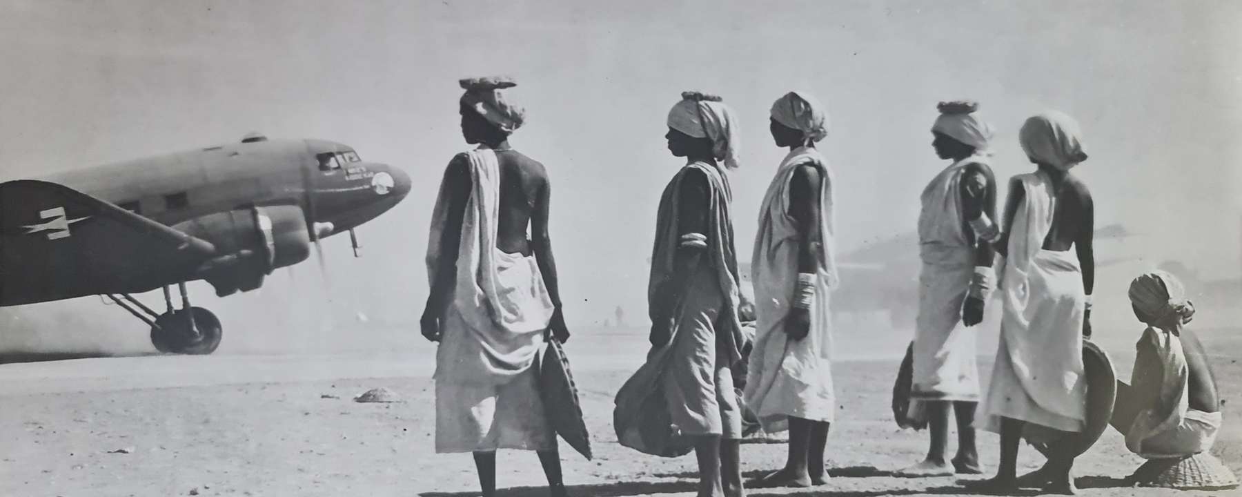 Black and white photograph of a group of six Manipuri women standing at a distance from military aircraft on a dusty airfield. They are holding baskets, and have their hair tied in wraps. Some have caps that enable them to carry loads on their heads.
