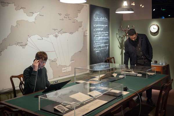 Two people look at files on a large table. Behind them is a map of the English Channel.