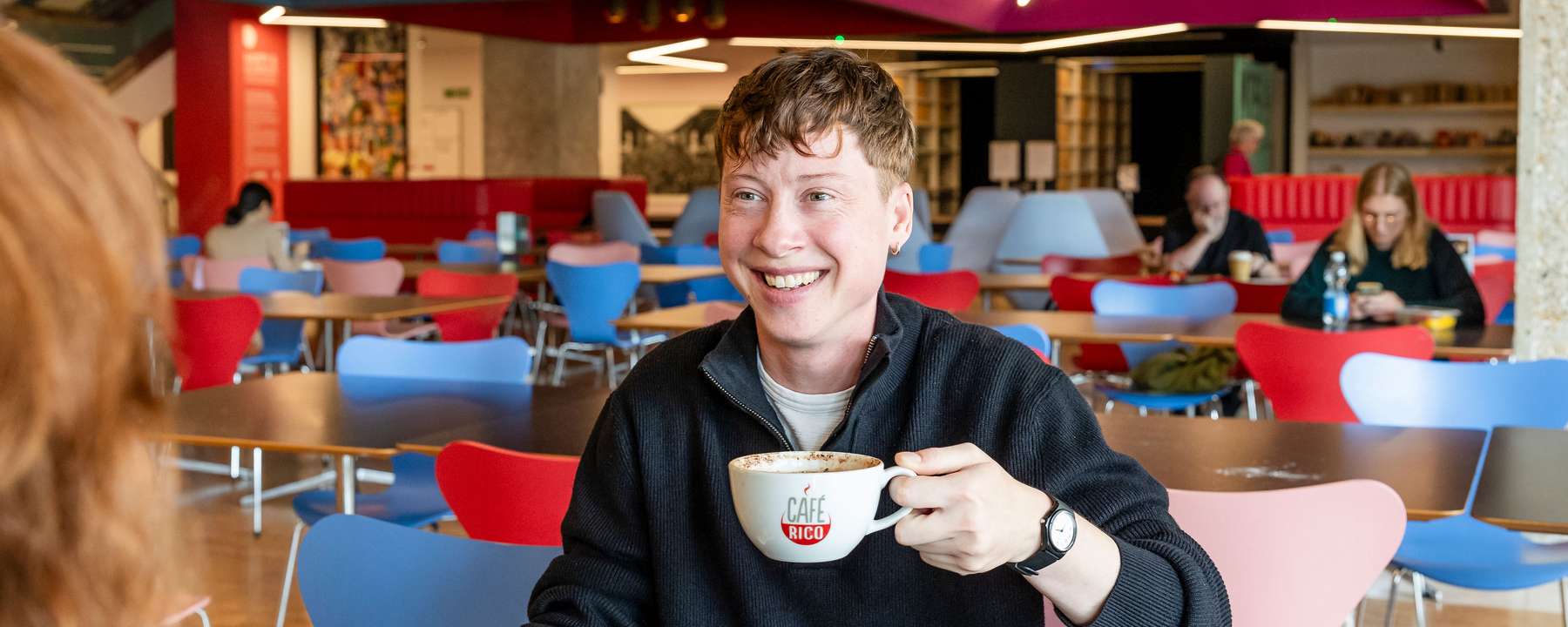 A smiling visitor seated at a table holds a cup of coffee in The National Archives café.