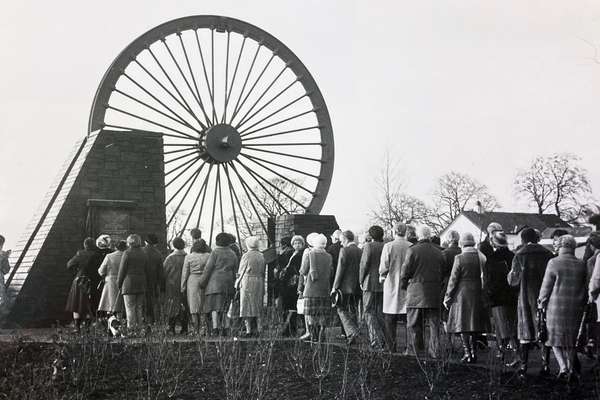A large wheel from a coal pit is above ground as a memorial. Tens of people are walking towards it.