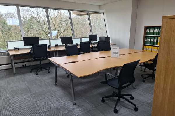 A bank of four desks joined together sits in front of three computers in a row, all with wheeled chairs beside, in front of a large window onto trees outside.