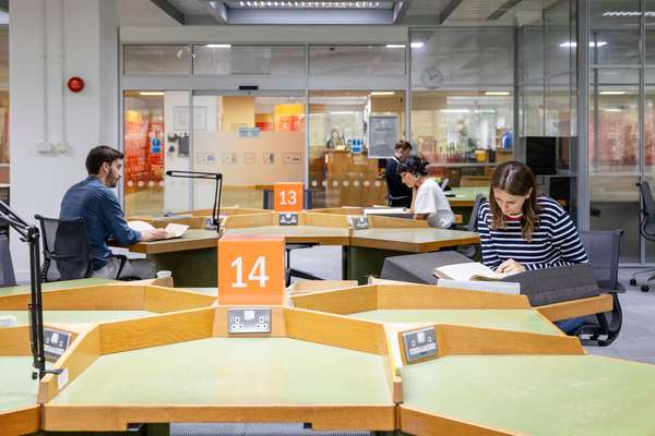 Four visitors sit at tables in a large, open room. Each is looking at a document.