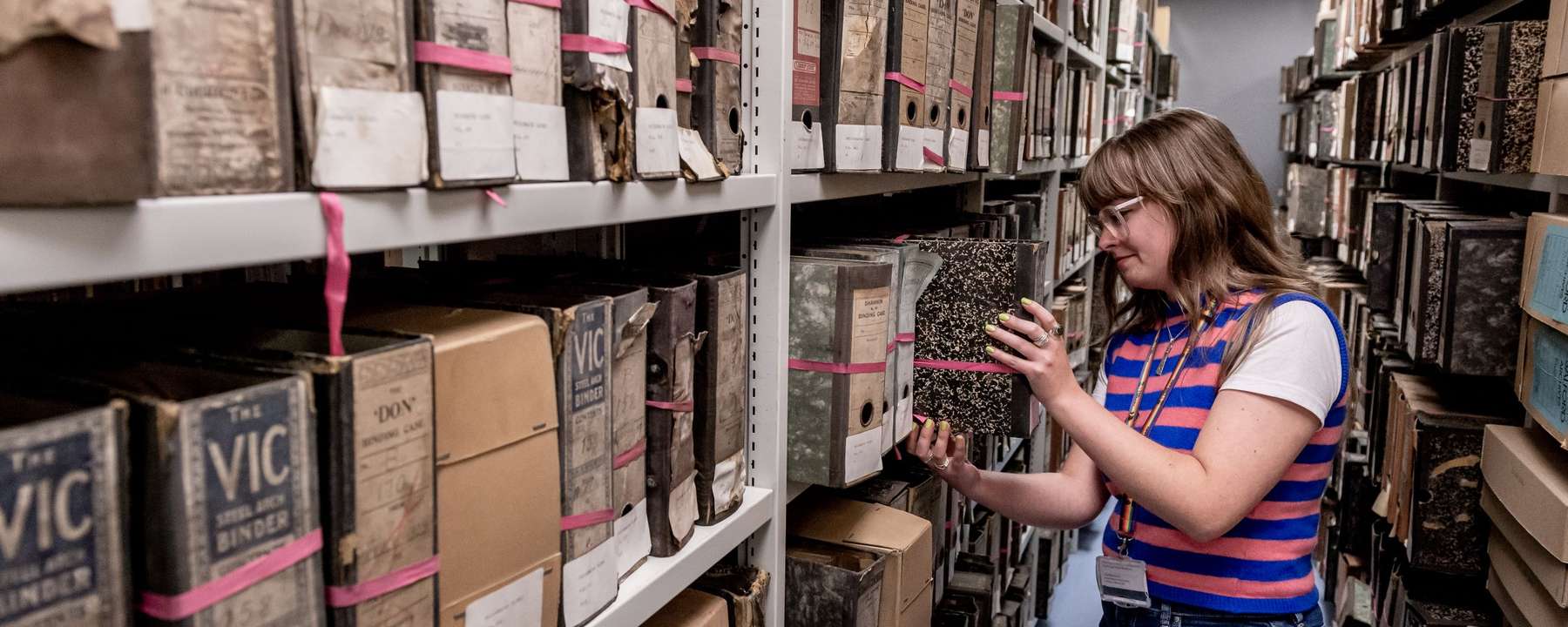A woman with long brown hair and glasses is standing in a repository corridor, in the middle of placing a hardbacked folder back on a shelf. All the shelves in the corridor are full of similar folders.