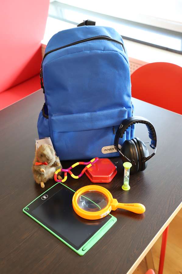 A small blue rucksack above a furry rat soft toy wearing a bow, a small sand timer, a pair of ear defenders, a flat tablet, a magnifying glass and some other toys.
