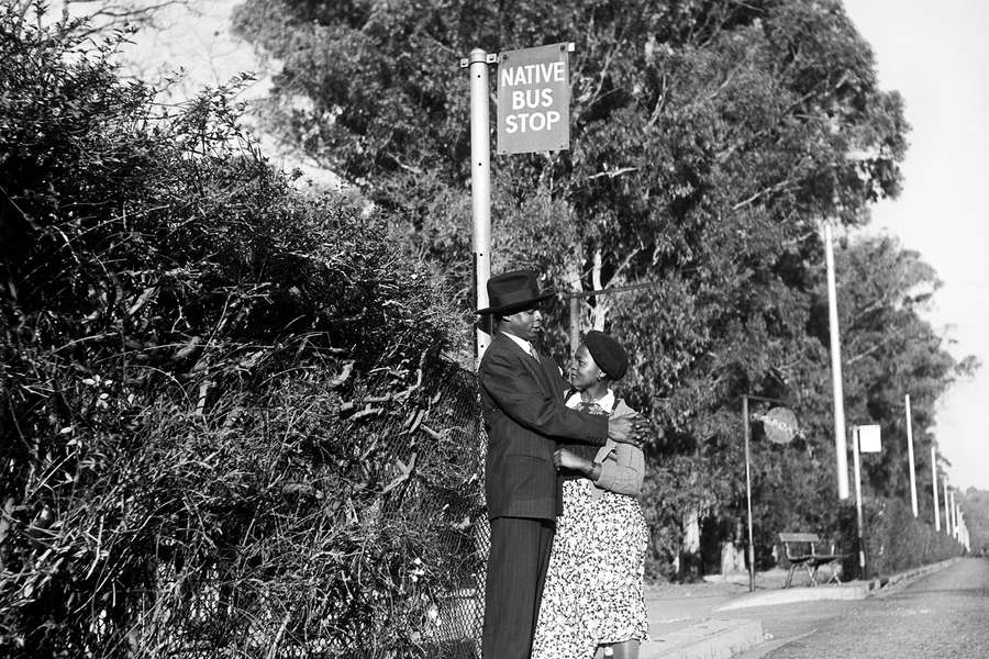 Black and white photography of a smartly-dressed Black couple hugging below a sign saying 'Native bus stop'.
