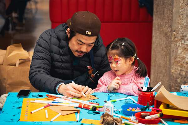 An adult sketches on a piece of card while a child with their face painted watches intently.