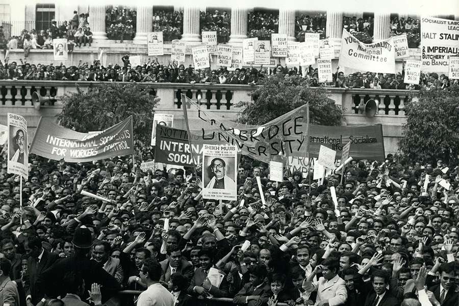 Black and white photograph of a large crowd of majority South Asian people. A large banner reading 'Cambridge University' is visible, alongside banners for various socialist movements and smaller placards featuring Sheikh Mujibur Rahman.