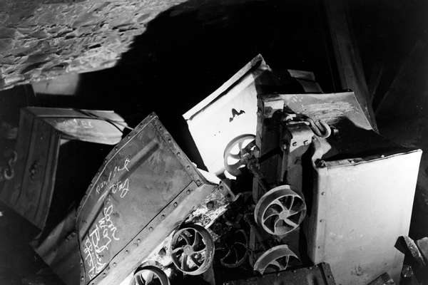 A photograph of inside a mine. There are coal trucks and rails upended. A torch light shines on them