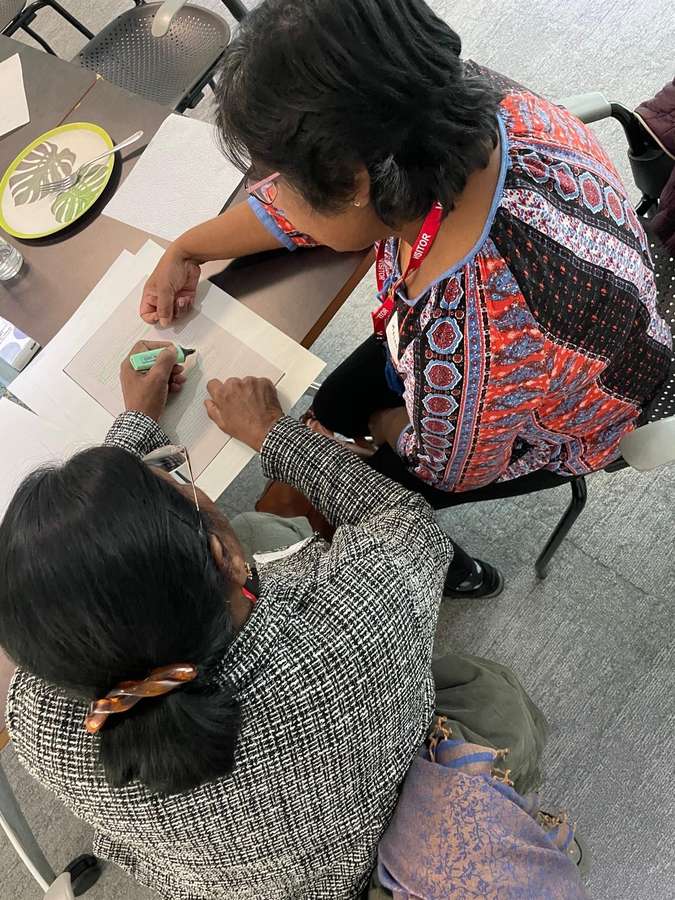 Two seated women hold a copy of an archival document in place while one of them highlights lines with a green marker pen.