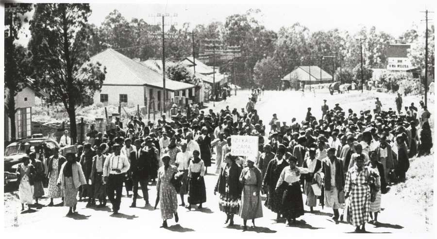 Black and white photograph showing a crowd of many dozens of women marching through a town square. One is holding a sign.