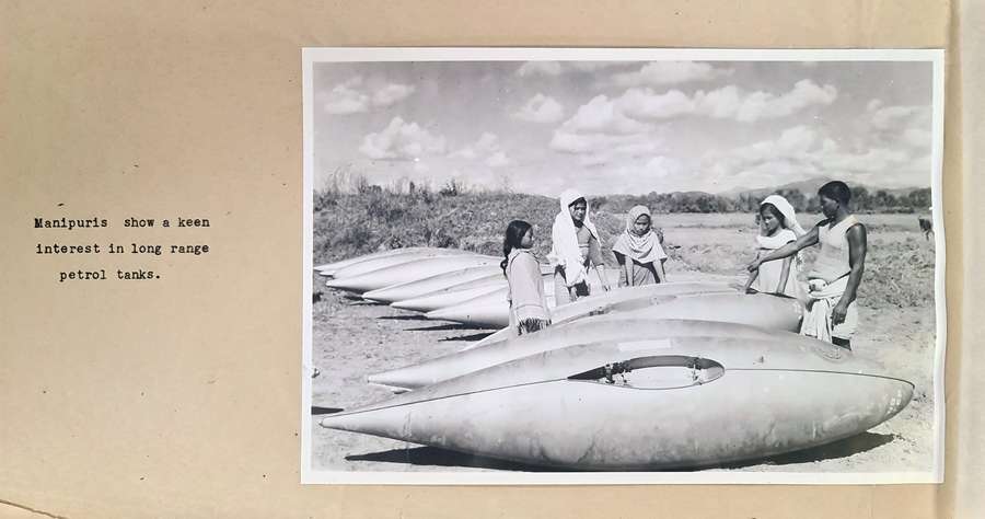 A typewritten caption beside a black and white photograph of four very young Manipuri women in various dresses (and most with loose head-coverings) standing in a large open space with mountains behind, around aeroplane petrol tanks.