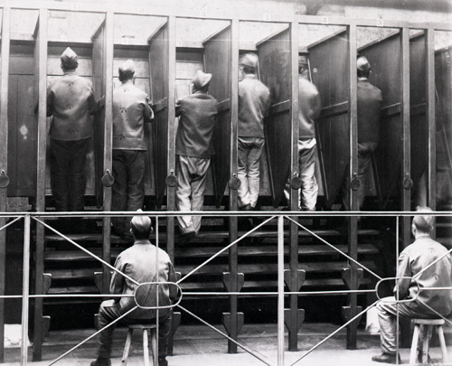 Prisoners walk on large wheels, separated by wooden dividers. Other prisoners sit on chairs waiting for their turn.