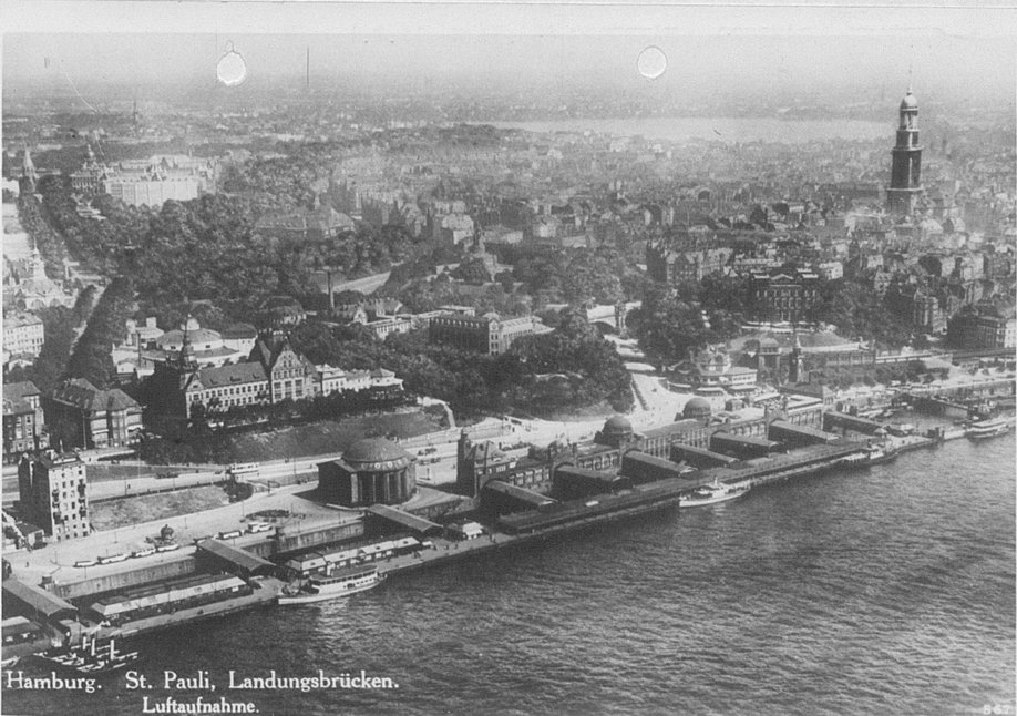 Monochrome photograph of a cityscape next to a body of water with large houses and ports, wide roads, and a tower visible.