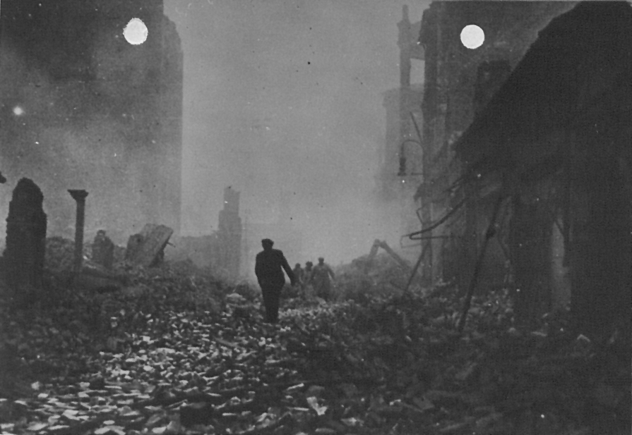 Monochrome photograph of a street filled with rubble and ruined houses, with a few people making their way through it.