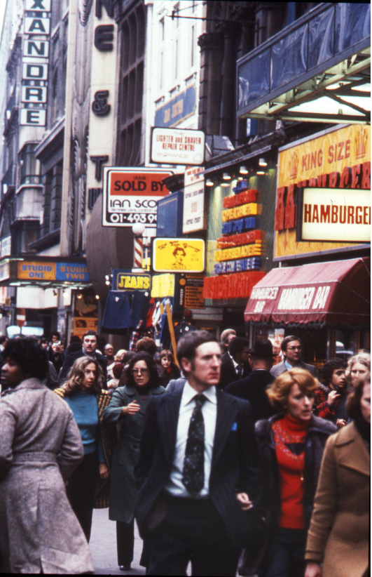 People walk down a bustling city street below a number of signs advertising various businesses.
