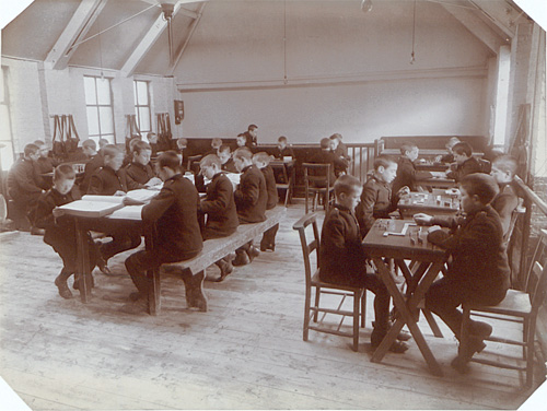 Classroom where boys sit at tables and play dominoes and read. The walls are mostly blank except for hanging bags.