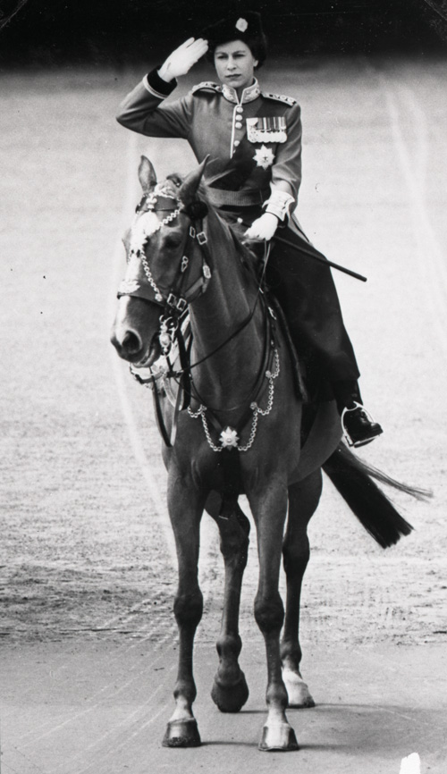 Monochrome photograph of a saluting woman in royal uniform riding on a horse.