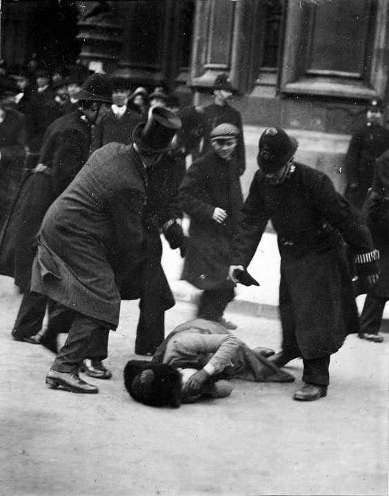 A women lies on the floor covering her face, at Black Friday demonstration in November 1910; she is surrounded by four men