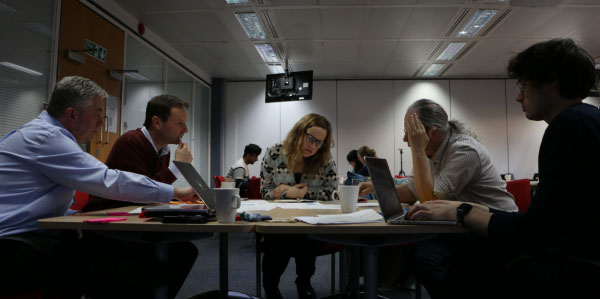 Five people sit round a table with laptops and papers on the table, looking down at some of the papers. There are three more people at another table in the background.