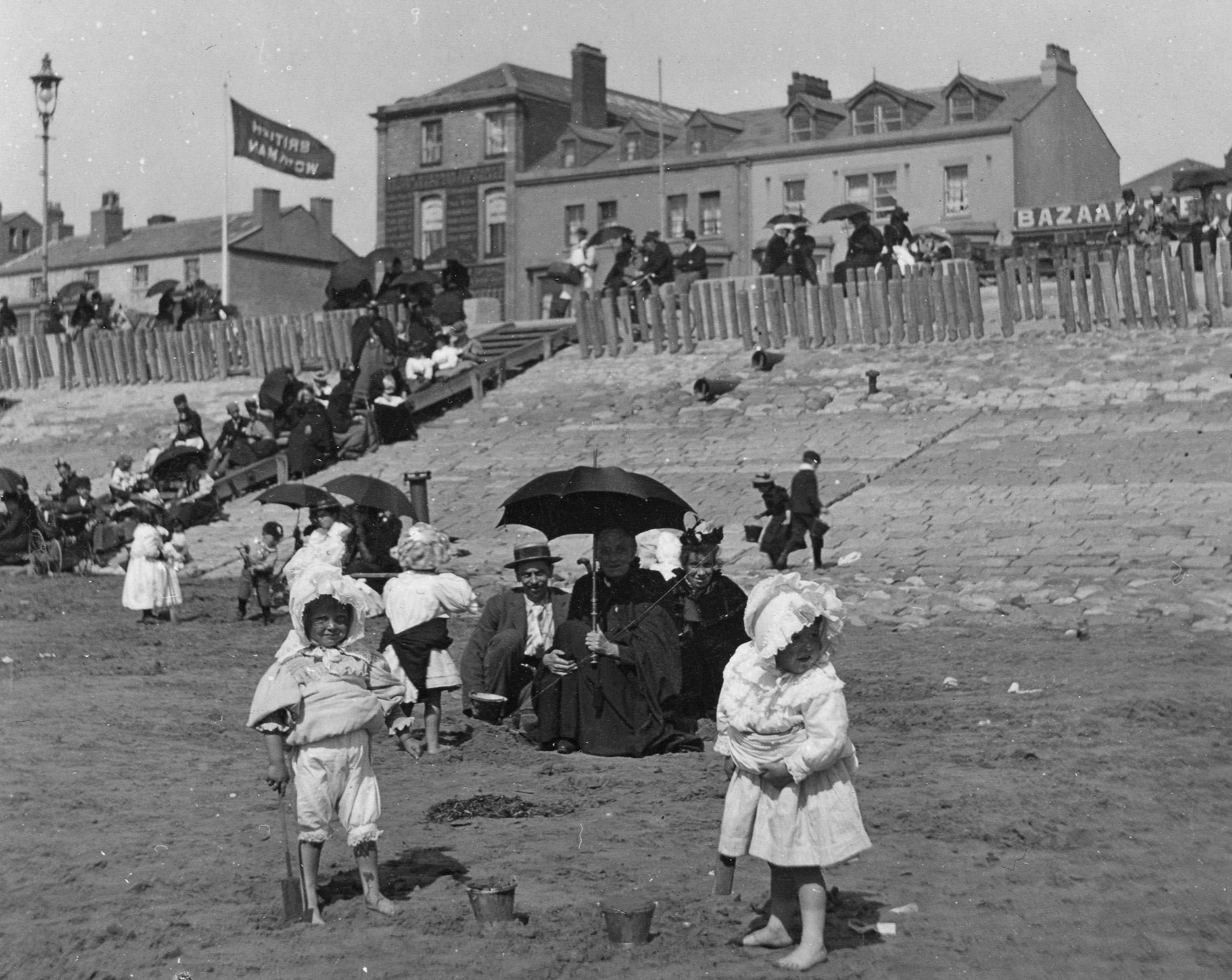 Two children in Victorian clothing stand on a sandy beach. A group of adults, one holding an umbrella, sit behind them and watch.