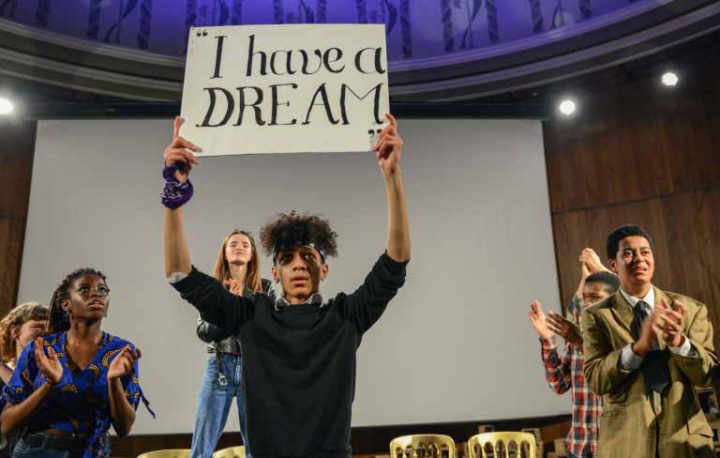 A performer on a stage holds a placard saying 'I have a dream'. The surrounding performers look towards the placard and applaud.