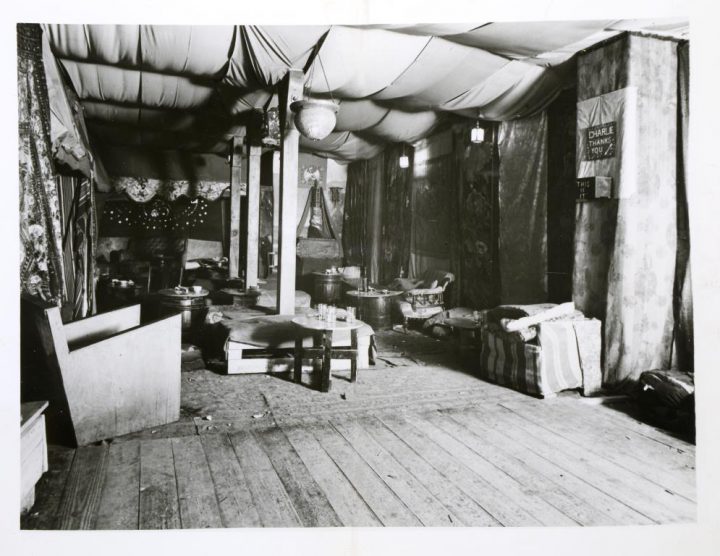 Monochrome photograph of the interior of a room with wooden foots, multiple chairs and tables, and decorated with draped fabric.