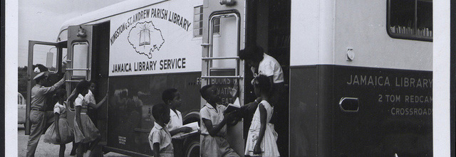 Detail of a monochrome photograph of children standing outside of a bus with the words 'Kingston & St. Andrew Parish Library Jamaica Library Service' on its side. The children are talking to adults standing inside the bus and two of the children hold books.