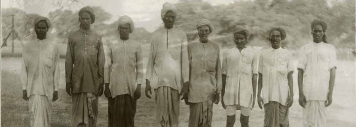 Monochrome photograph of eight Sikh men standing in a row and looking at the camera.