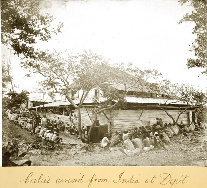 Monochrome photograph of a building with group of people sitting around it. A handwritten caption underneath says 'Coolies arrived from India at Depot'.