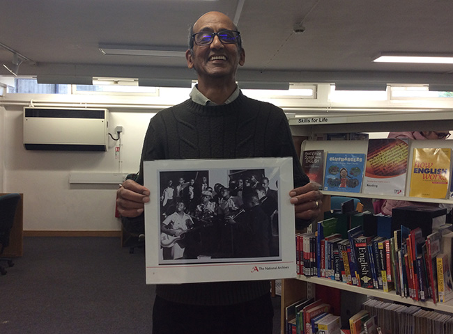 A smiling man holds up a laminated monochrome photograph of a scene of people playing music in the Bechuanaland Protectorate (now Botswana).