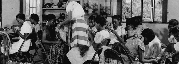 Detail of a monochrome photograph of a group of Jamaican women sitting on chairs inside a room next to piles of raffia. They are all making handicrafts using the raffia.