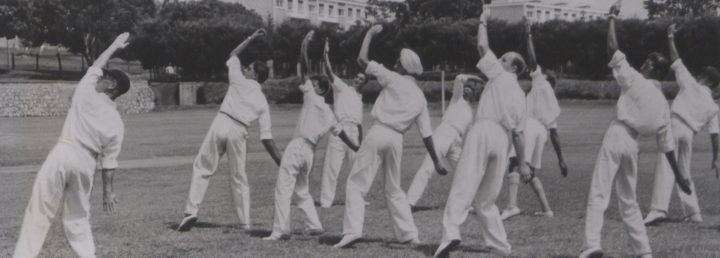 Monochrome photograph of men dressed in white cricket clothes on a lawn stretching their arms in the air.