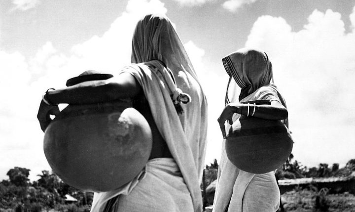 Two Bengali women viewed from behind, carrying large ceramic pots balanced on their hips.