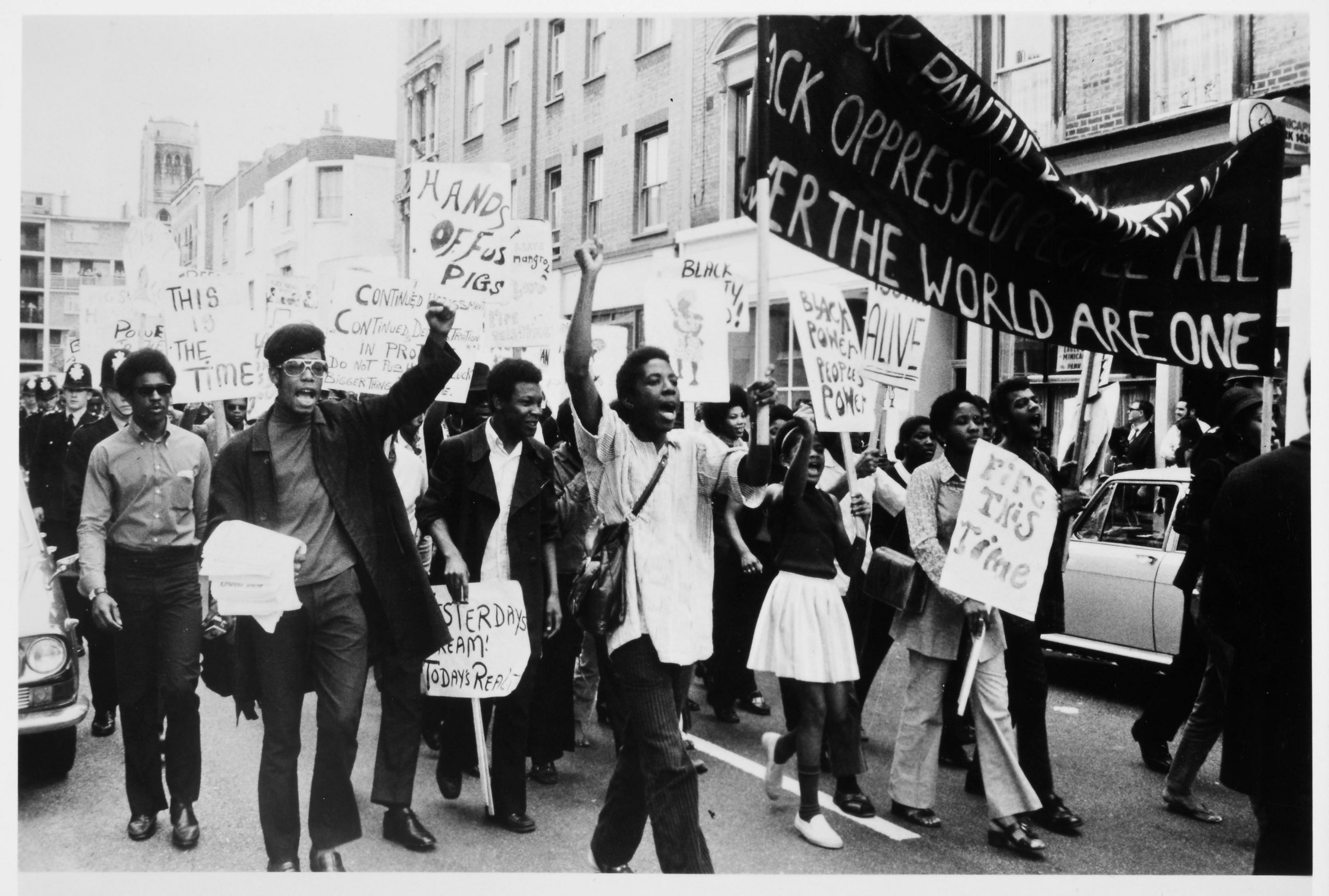 Monochrome photograph of a protest in which a group of Black British people walk forwards while holding placards with messages against police oppression. A group of police officers follow behind.