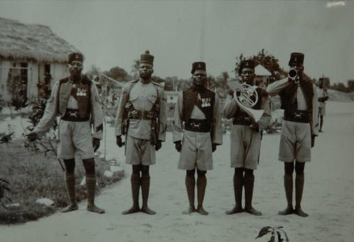 Five men in uniform stand in a row looking at the camera. The two men on the right are holding a French horn and trumpet.