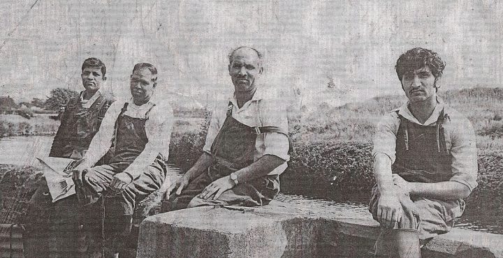 Monochrome photograph of four South Asian men in overalls sitting on a wall looking at the camera.