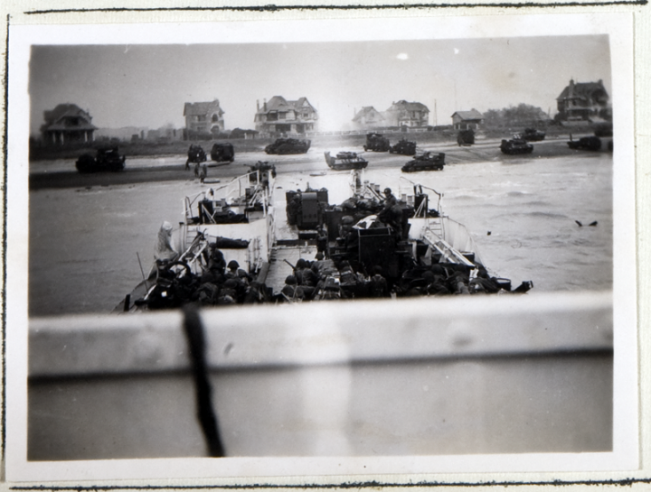 Operation Neptune landing craft during the D-Day Landings in June 1944 (catalogue reference ADM 199/1660). In the foreground are troops and landing craft readying to leave a boat approaching the shore; int he background are tanks and other vehicles on a beach in front of several detached houses.