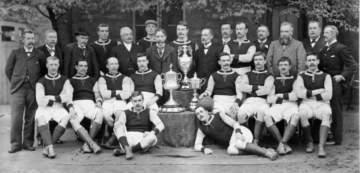 A cropped version of a photograph of Aston Villa Football Club, showing the whole team in club colours and ten older men in suits, gathered around three trophies. The photo was registered for copyright in 1896 (catalogue reference COPY 1/424/87).