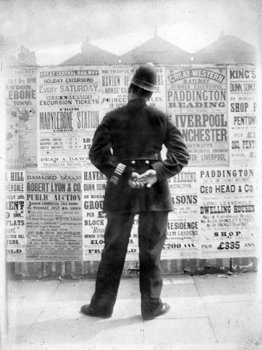 A rear view of a policeman stood in front of a wall of bill posters with his hands behind his back, apparently reading the posters.