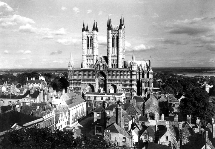 A wide shot of Lincoln Cathedral from the front and the surrounding landscape, with houses in the foreground and flat countryside in the background. The photo is from the 1930s (catalogue reference INF 9/699).