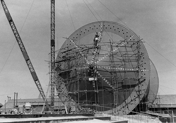 A giant metal cylinder covered in scaffolding supporting two workmen. This is the construction of the 'Conun' drum in 1943, used to lay pipe under the English Channel for the Pipe Line Under The Ocean (PLUTO) project (catalogue reference POWE 45/61).