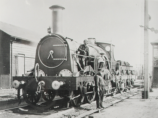 A broad gauge 'Lightning' locomotive of the Great Western Railway, stationary in a yard, 17 July 1889 (catalogue reference RAIL 1014/19). A man and a child are shown stood on the side of the locomotive and another man is stood on the ground in front.