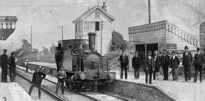 A locomotive engine stationary at Ludgershall train station, on the Midland and South Western Junction Railway, in 1901 (catalogue reference RAIL 1014/36/16). Fourteen men pose for the camera, stood side by side across the platform, on the tracks and on the locomotive itself.